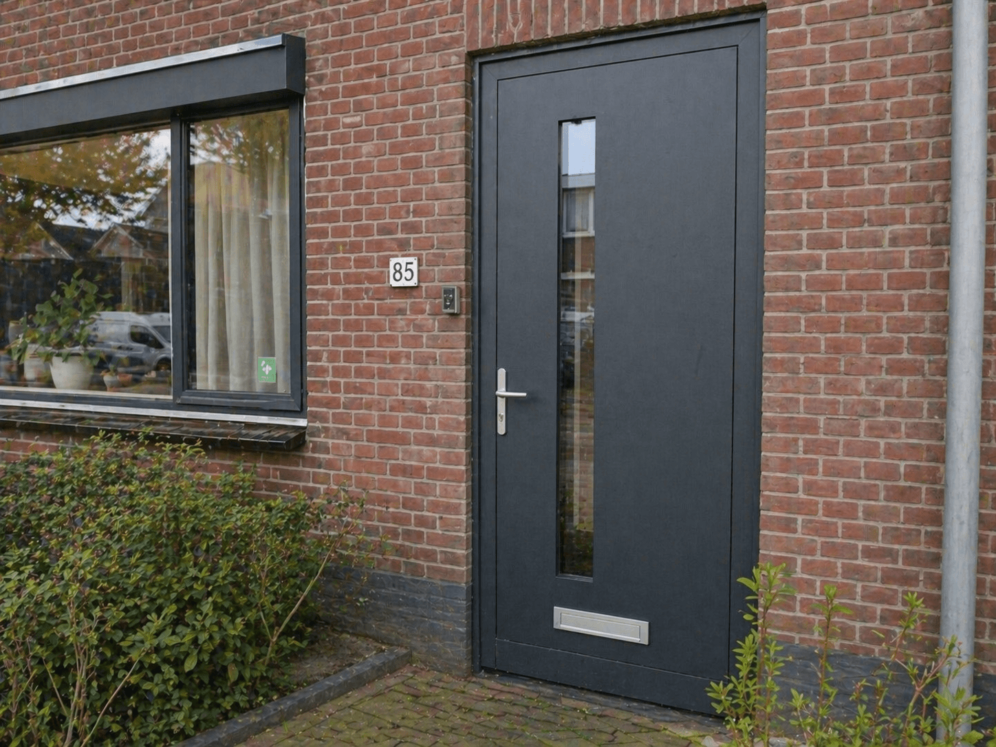 Antraciete kunststof voordeur met smalle glasstrook in rode bakstenen gevel